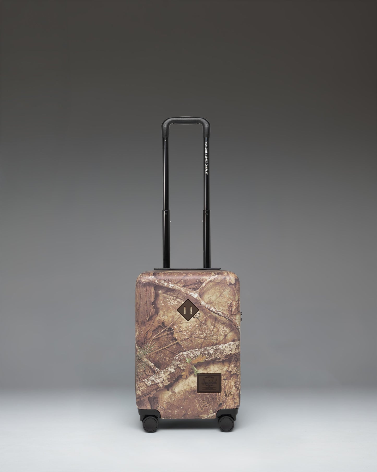 Herschel Heritage Carry On Luggage in Realtree APX Camo pattern, showing the front view with a black zipper and signature logo label.