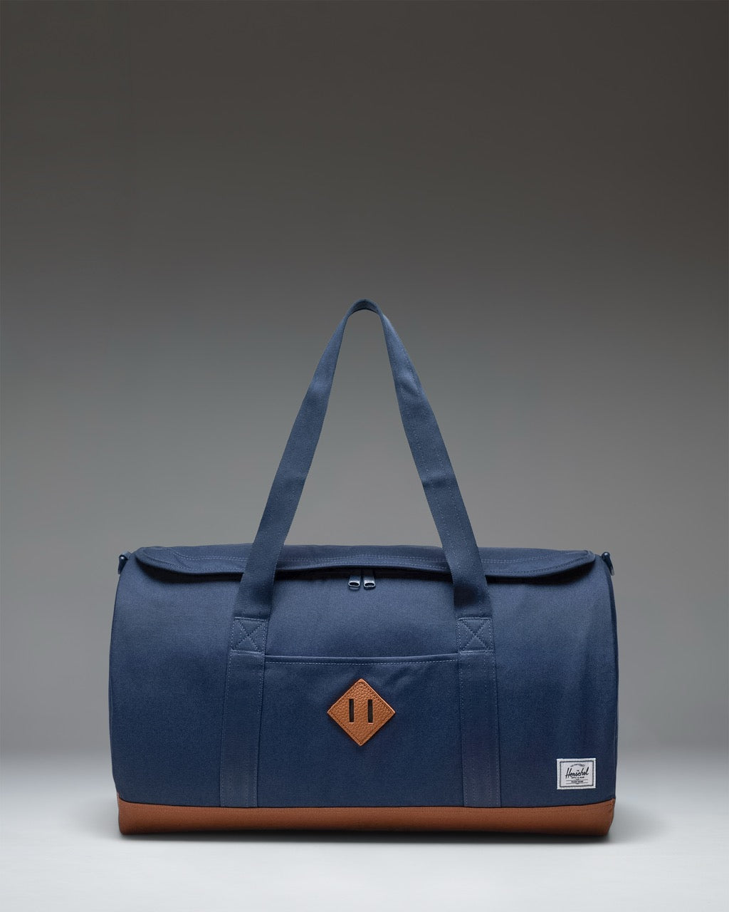 Herschel Heritage Duffle bag in Navy Blue with Saddle Brown handles and detailing, shown from a three-quarter angle on a white background.