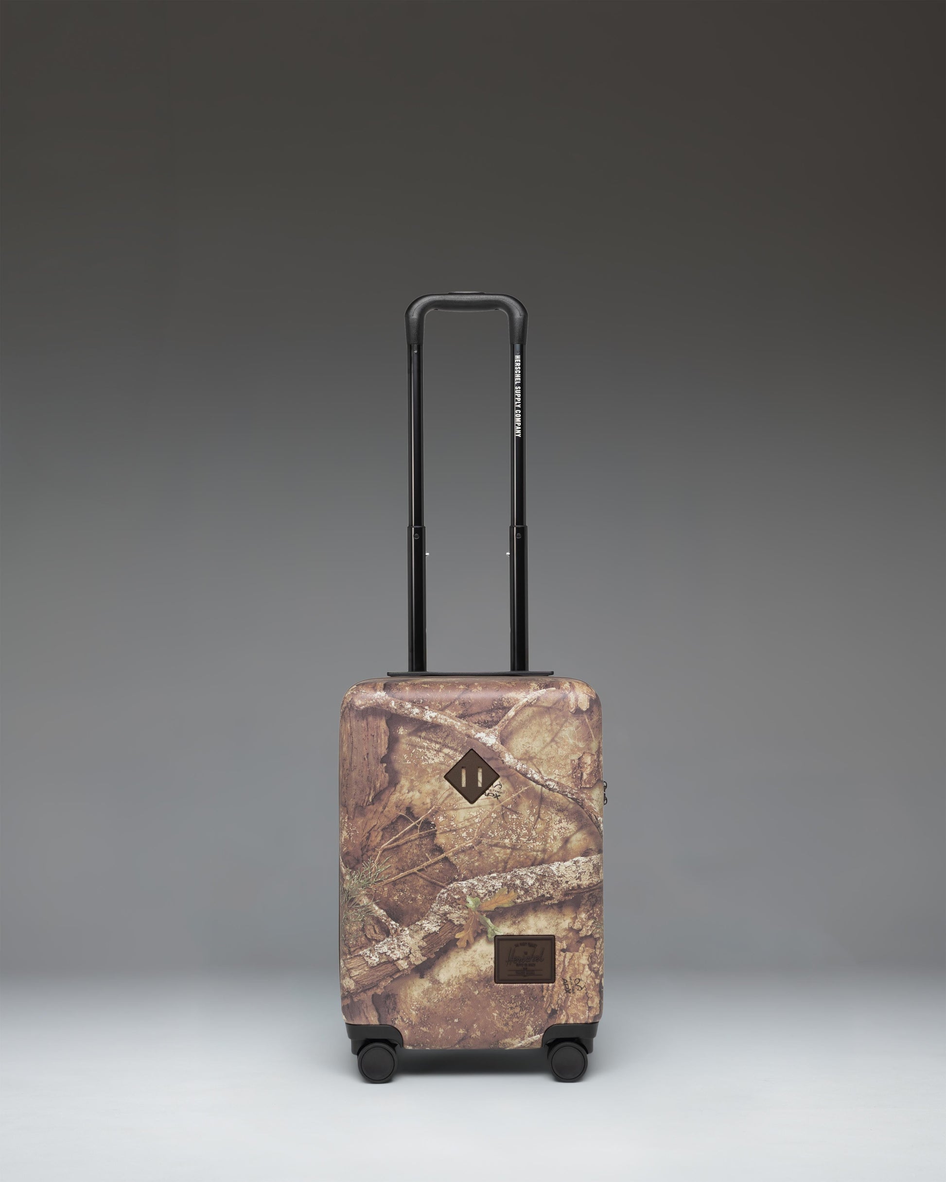 Herschel Heritage Carry On Luggage in Realtree APX Camo pattern, showing the front view with a black zipper and signature logo label.