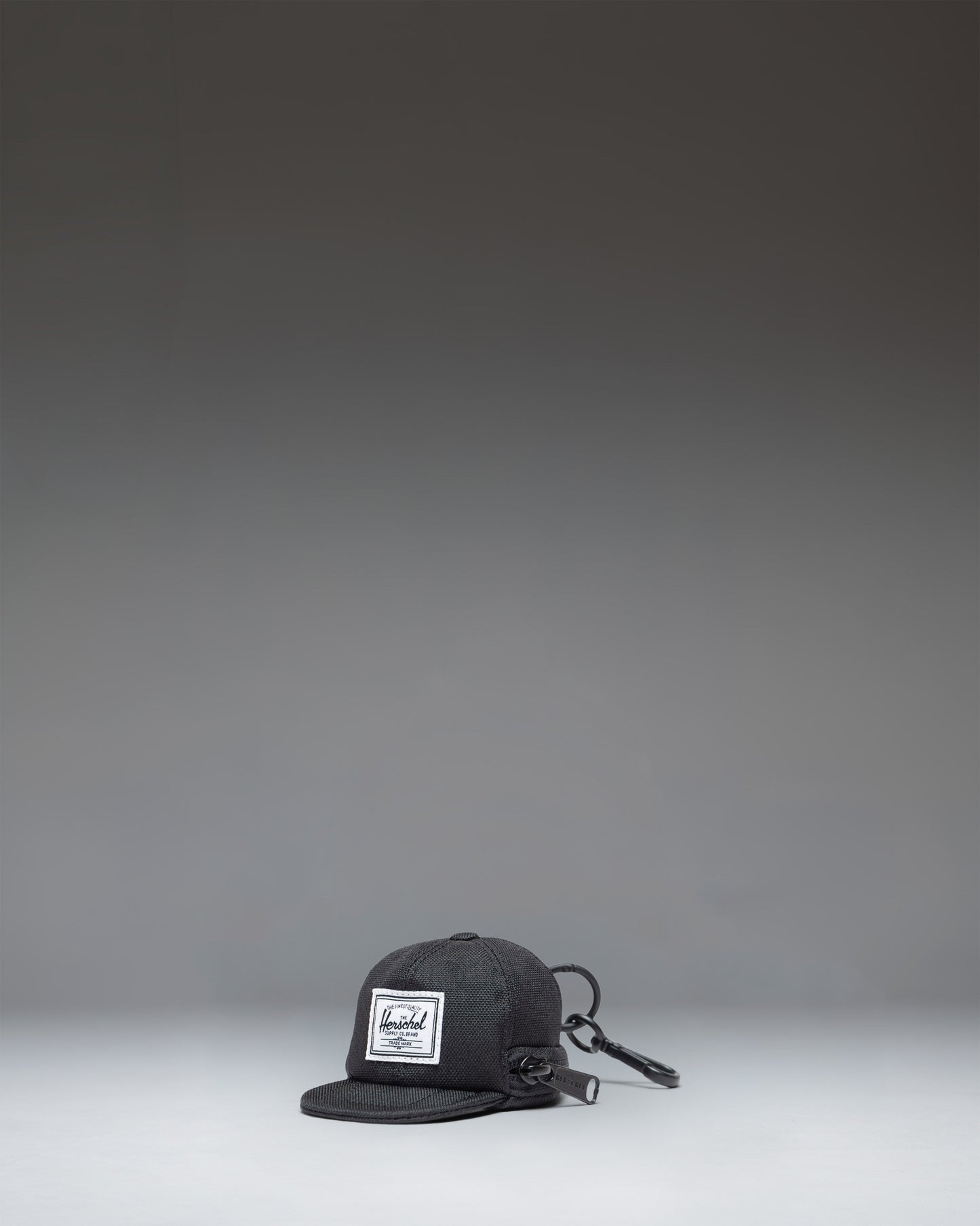 Herschel Supply Co. Sylas Classic Cap Bag Charm in black, showing the front view of a miniature baseball cap keychain accessory.
